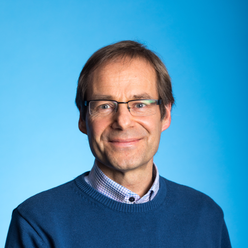 Headshot of Dirk Pesch, wearing a dark blue jumper over a blue and white check shirt and standing against a sky blue background.