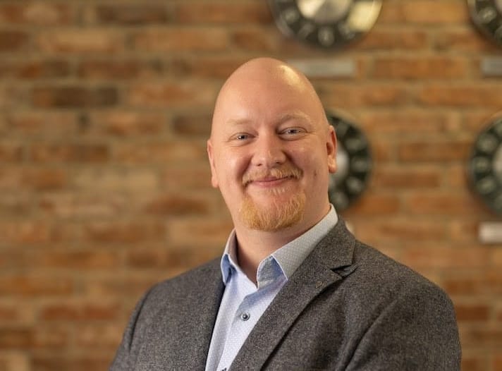 Headshot of Tom Deegan, smiling against a red brick wall.